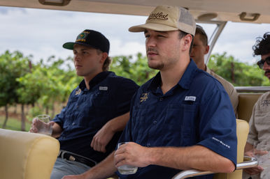 Group of young men in caps and navy shirts seated on an open-air vineyard shuttle, holding tasting glasses while passing rows of grapevines on a wine-country tour.