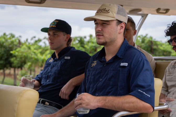Group of young men in caps and navy shirts seated on an open-air vineyard shuttle, holding tasting glasses while passing rows of grapevines on a wine-country tour.