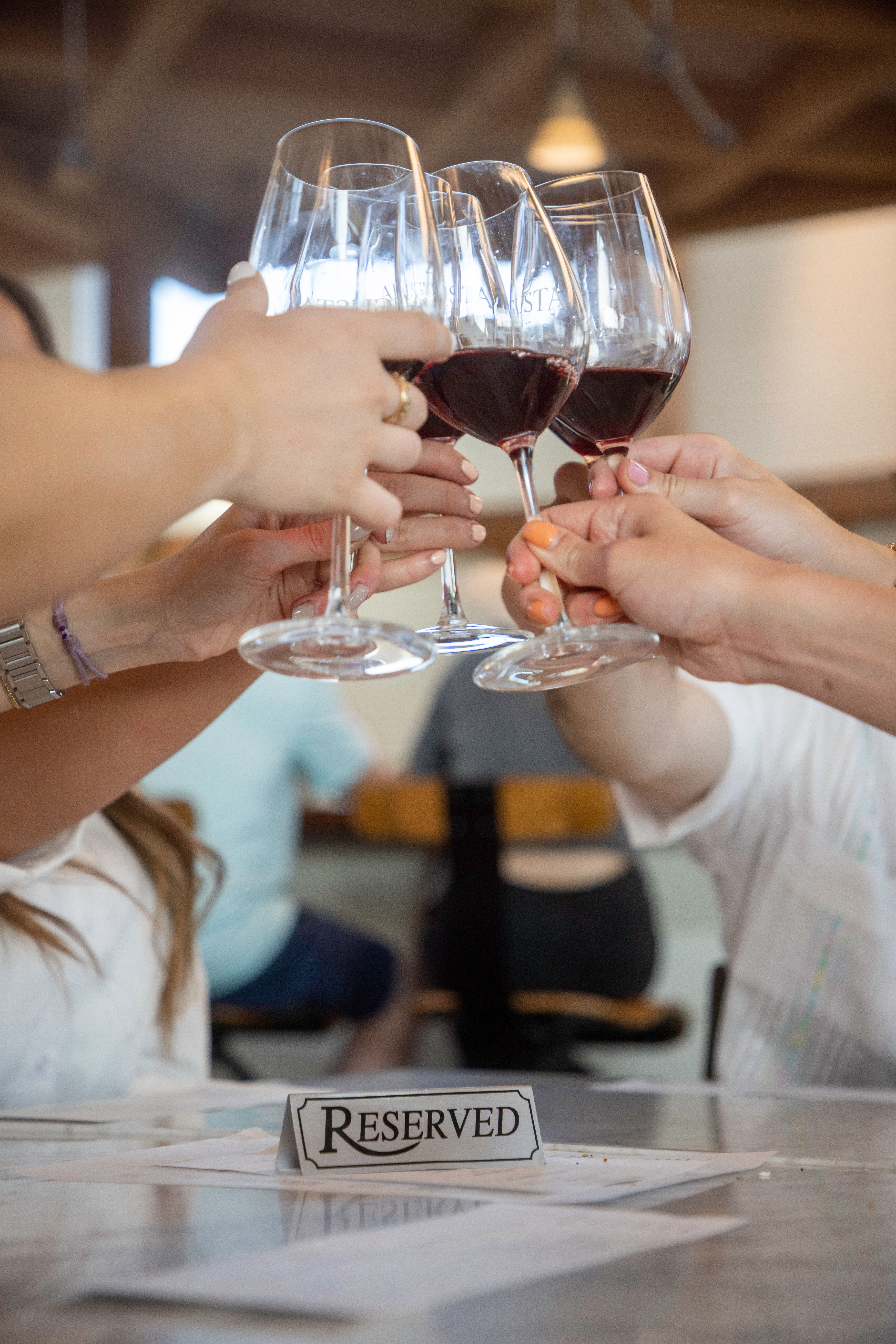 Group cheers with red wine glasses clinking above a 'Reserved' sign on a restaurant table — casual wine-tasting vibe.