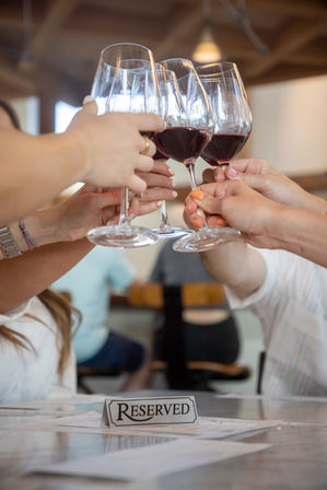 Group cheers with red wine glasses clinking above a 'Reserved' sign on a restaurant table — casual wine-tasting vibe.