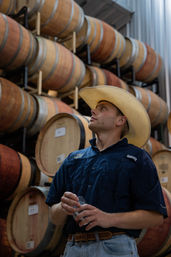 Man in a cowboy hat holding a tasting glass in a winery barrel room, surrounded by stacked oak wine barrels in a rustic cellar
