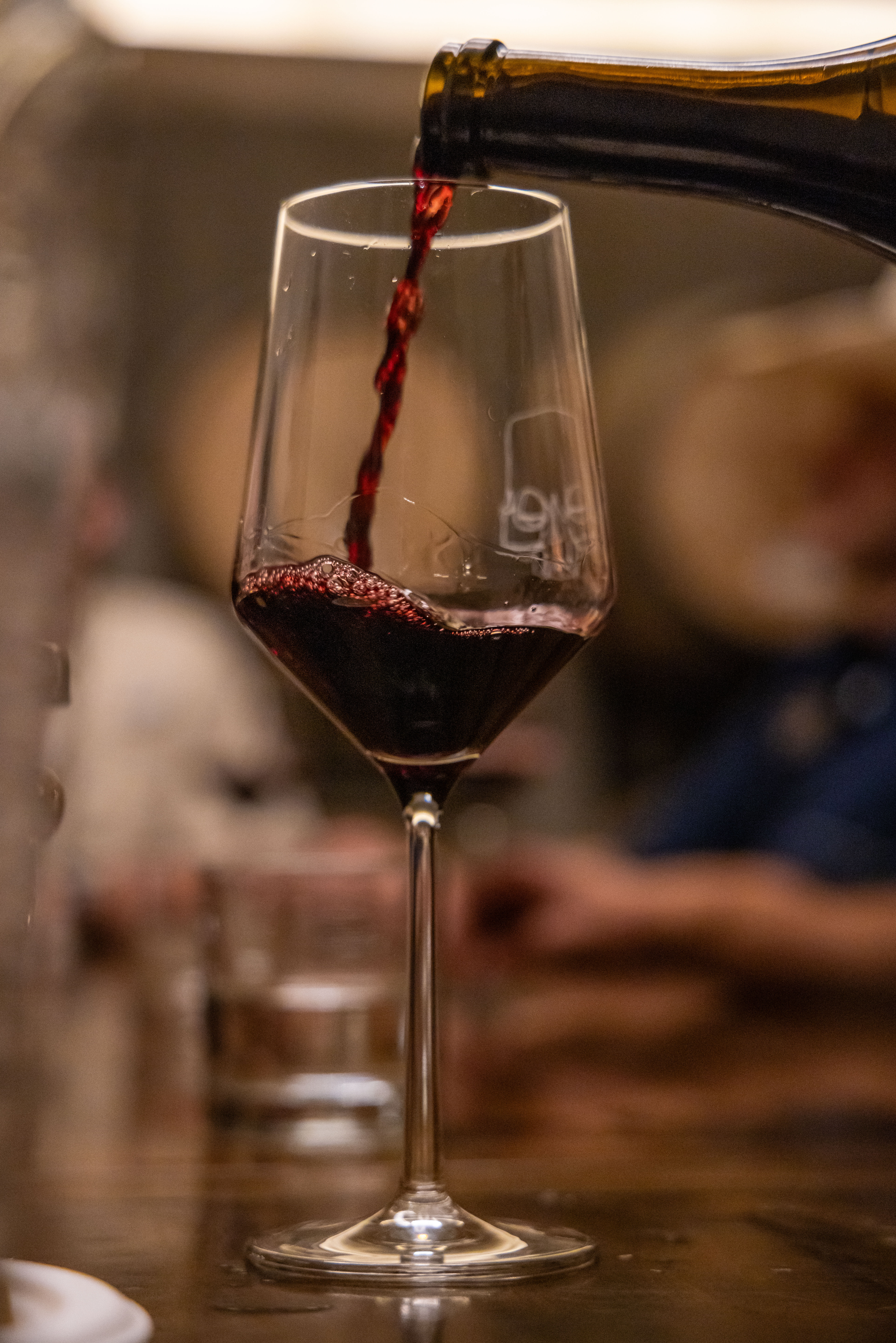 Close-up of deep red wine being poured from a bottle into a tall stemmed glass on a dimly lit restaurant table, cozy wine bar ambiance