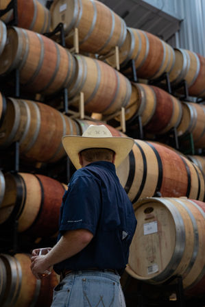 Person in a cowboy hat and jeans holding a wine glass in a winery barrel room with rows of stacked oak wine barrels.