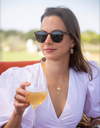Woman in a white blouse and tortoiseshell sunglasses sipping chilled white wine on a sunny outdoor patio, wearing gold necklaces and earrings