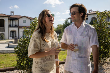 Two friends wearing sunglasses laugh and sip white wine in a sunny Mediterranean-style courtyard with stucco villas and lush greenery.