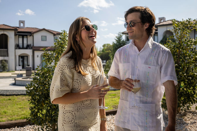 Two friends wearing sunglasses laugh and sip white wine in a sunny Mediterranean-style courtyard with stucco villas and lush greenery.