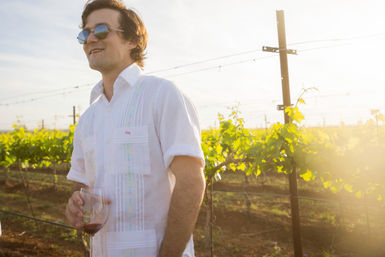 Smiling person in a white linen shirt and sunglasses holding a glass of red wine in a sunlit vineyard at golden hour with rows of green grapevines and trellises