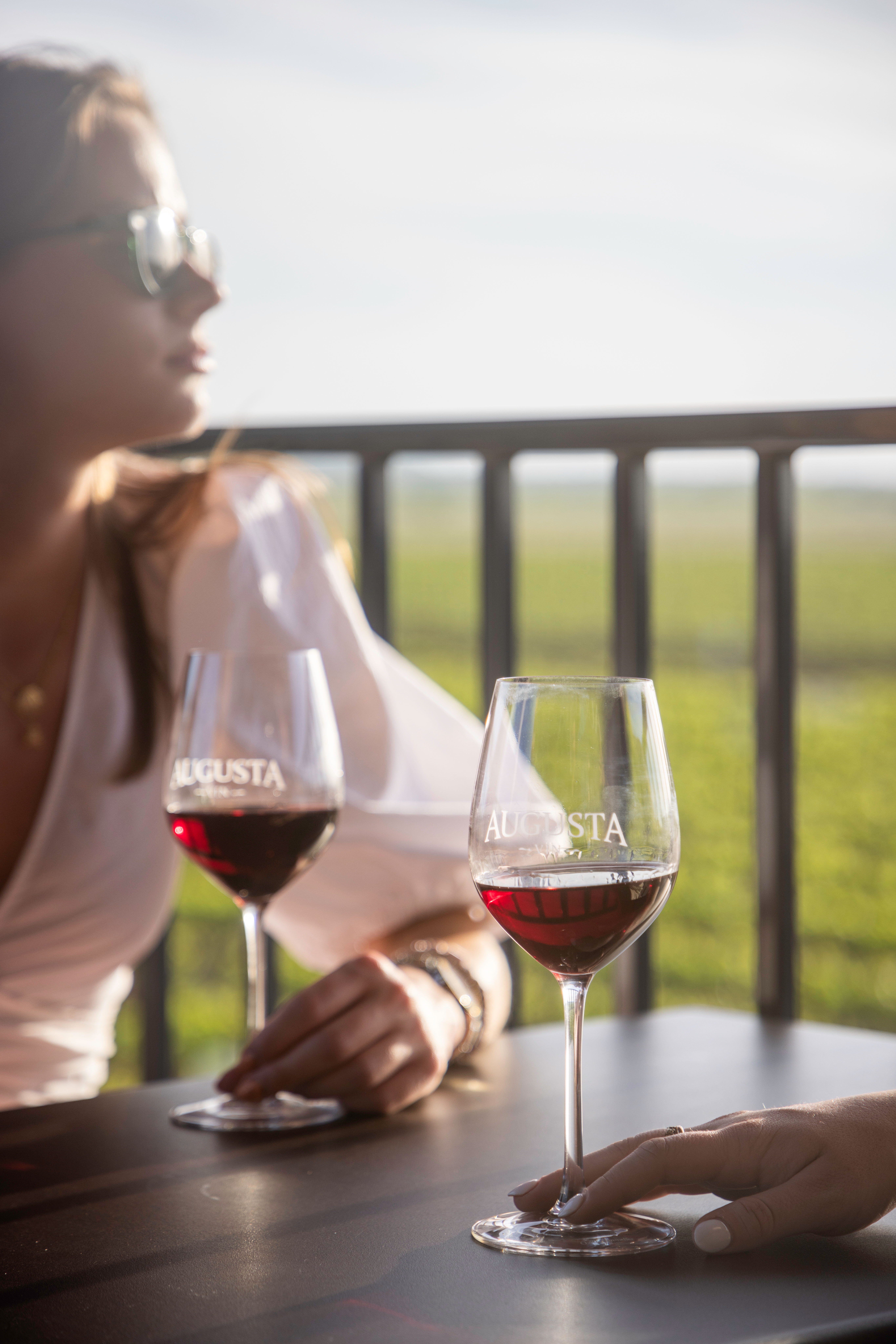Sunlit vineyard terrace with two red wine glasses on a table, hands resting on the stems and a blurred person in sunglasses enjoying an outdoor wine tasting.