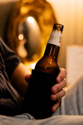 Close-up of a hand holding a chilled brown beer bottle in a black koozie on a couch, warm ambient lighting and a decorative mirror softly blurred in the cozy living room background.