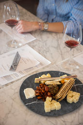 Assorted cheese and cracker board on a round slate—cubed cheeses, goat cheese, roasted almonds, crackers and tapenade—paired with two glasses of red wine on a marble table.