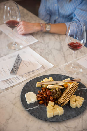 Assorted cheese and cracker board on a round slate—cubed cheeses, goat cheese, roasted almonds, crackers and tapenade—paired with two glasses of red wine on a marble table.