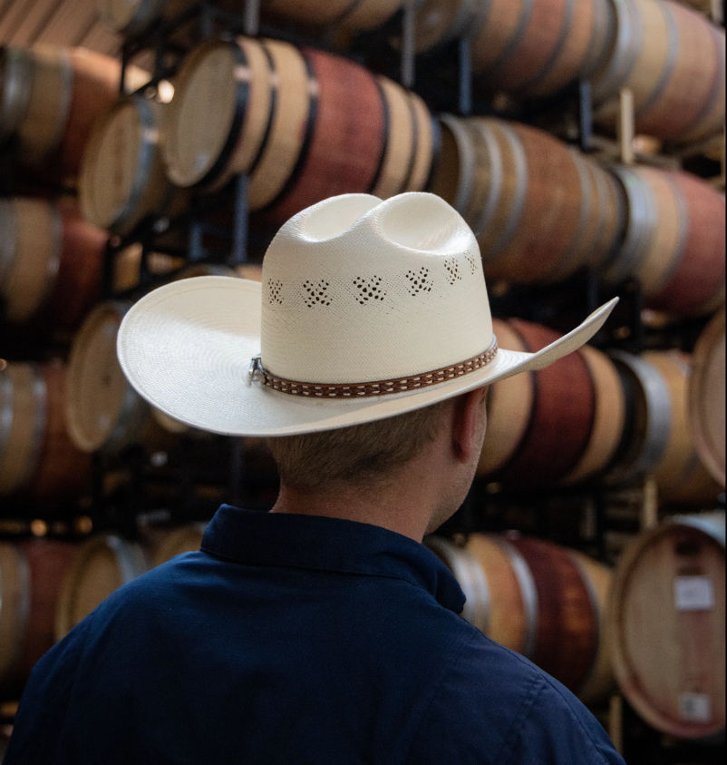 Back view of a person wearing a white cowboy hat in a winery barrel room, surrounded by rows of stacked oak wine barrels.
