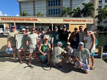 Group of men in casual summer clothes and matching tiki-themed shirts posing on a sunny marina dock in front of a charter boat canopy, with palm trees and waterfront buildings in the background.