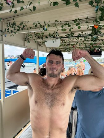Shirtless man playfully flexing his biceps aboard a vine-decorated party boat, with sunbathers, other boats, and water in the background