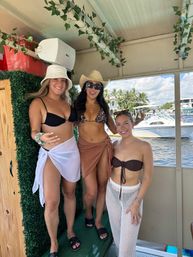 Three women in bikinis and sun hats smiling on a covered pontoon boat, with palm trees, yachts and a sunny marina in the background.