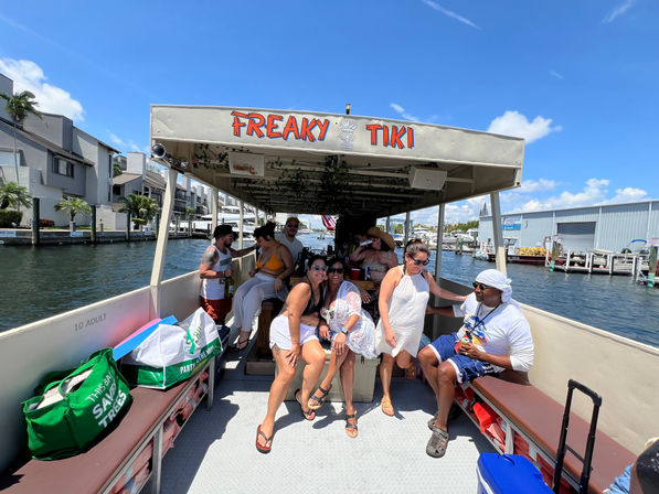 Tiki-style party boat cruising a sunny marina canal, friends laughing with drinks under a covered deck, docks and yachts visible