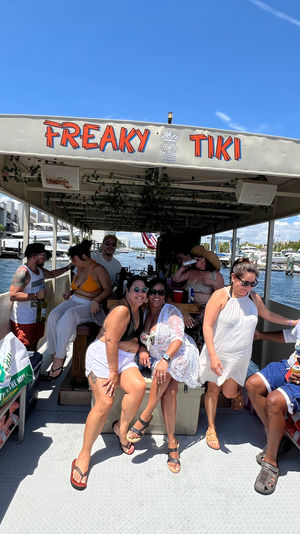 Tiki-style party boat cruising a sunny marina canal, friends laughing with drinks under a covered deck, docks and yachts visible