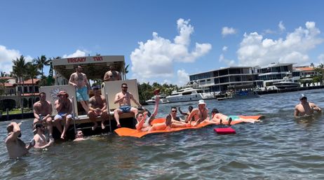 Group of people enjoying a sunny day on a floating tiki bar and orange inflatable mat in a marina-lined canal with yachts and waterfront homes.