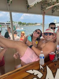 Boat party scene with friends lounging under a shaded canopy on a sunny Miami waterfront, palm trees, boats and skyline in the background, sunglasses, drinks and a pink bucket hat.