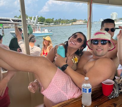 Boat party scene with friends lounging under a shaded canopy on a sunny Miami waterfront, palm trees, boats and skyline in the background, sunglasses, drinks and a pink bucket hat.