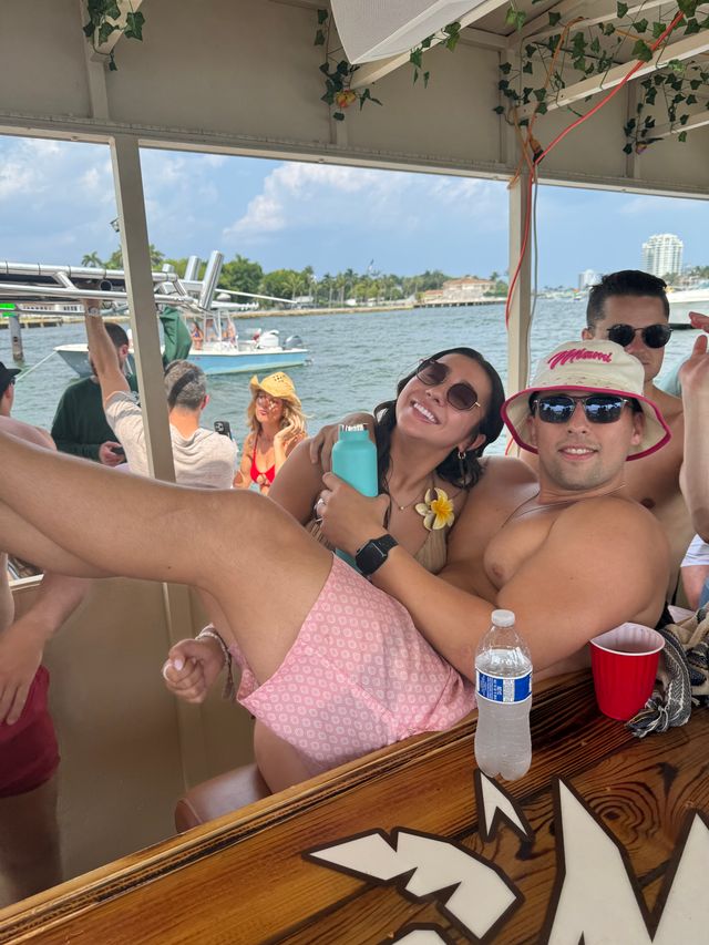 Boat party scene with friends lounging under a shaded canopy on a sunny Miami waterfront, palm trees, boats and skyline in the background, sunglasses, drinks and a pink bucket hat.