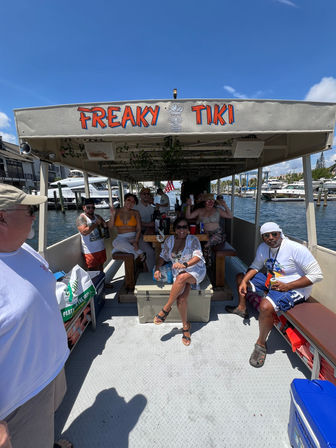 Smiling group enjoying a tiki‑themed party boat under a shaded canopy on a sunny day, cruising a marina with yachts docked nearby.