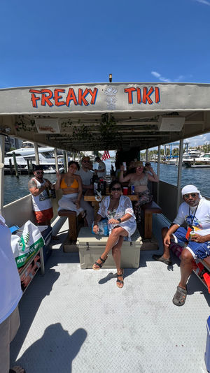 Smiling group enjoying a tiki‑themed party boat under a shaded canopy on a sunny day, cruising a marina with yachts docked nearby.