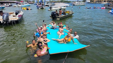 Sunny boat party at a busy marina — a group relaxing on a bright blue floating mat and in the water, surrounded by boats, inflatables, and swimmers.