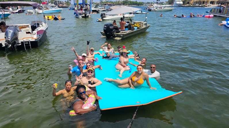 Sunny boat party at a busy marina — a group relaxing on a bright blue floating mat and in the water, surrounded by boats, inflatables, and swimmers.