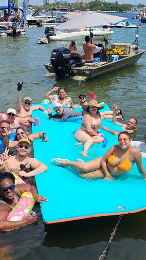 Sunny boat party at a busy marina — a group relaxing on a bright blue floating mat and in the water, surrounded by boats, inflatables, and swimmers.