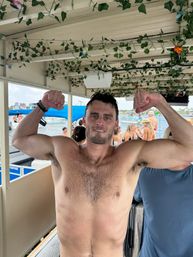Shirtless man flexes biceps on a lively party boat beneath a vine-decorated canopy, with people in swimsuits and boats on a sunny marina waterway.