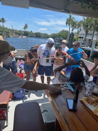Group of adults playing limbo with shot cups during a sunny boat party on a palm‑lined waterfront canal
