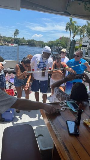 Group of adults playing limbo with shot cups during a sunny boat party on a palm‑lined waterfront canal