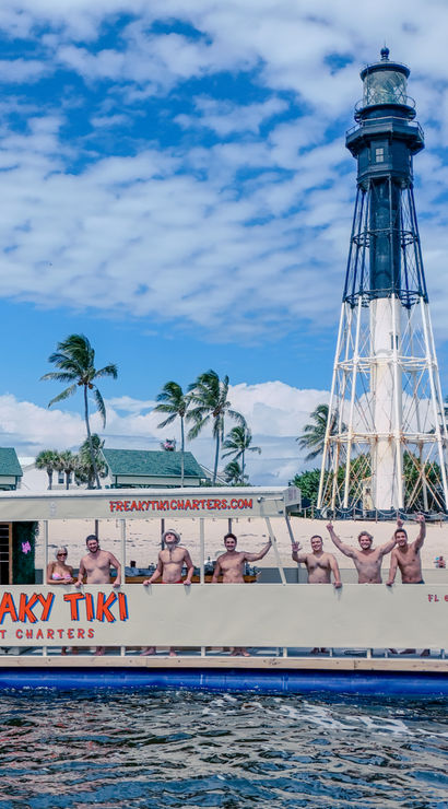 Party tiki boat with people waving near a tall black-and-white lighthouse off a palm-lined Florida beach under bright blue skies