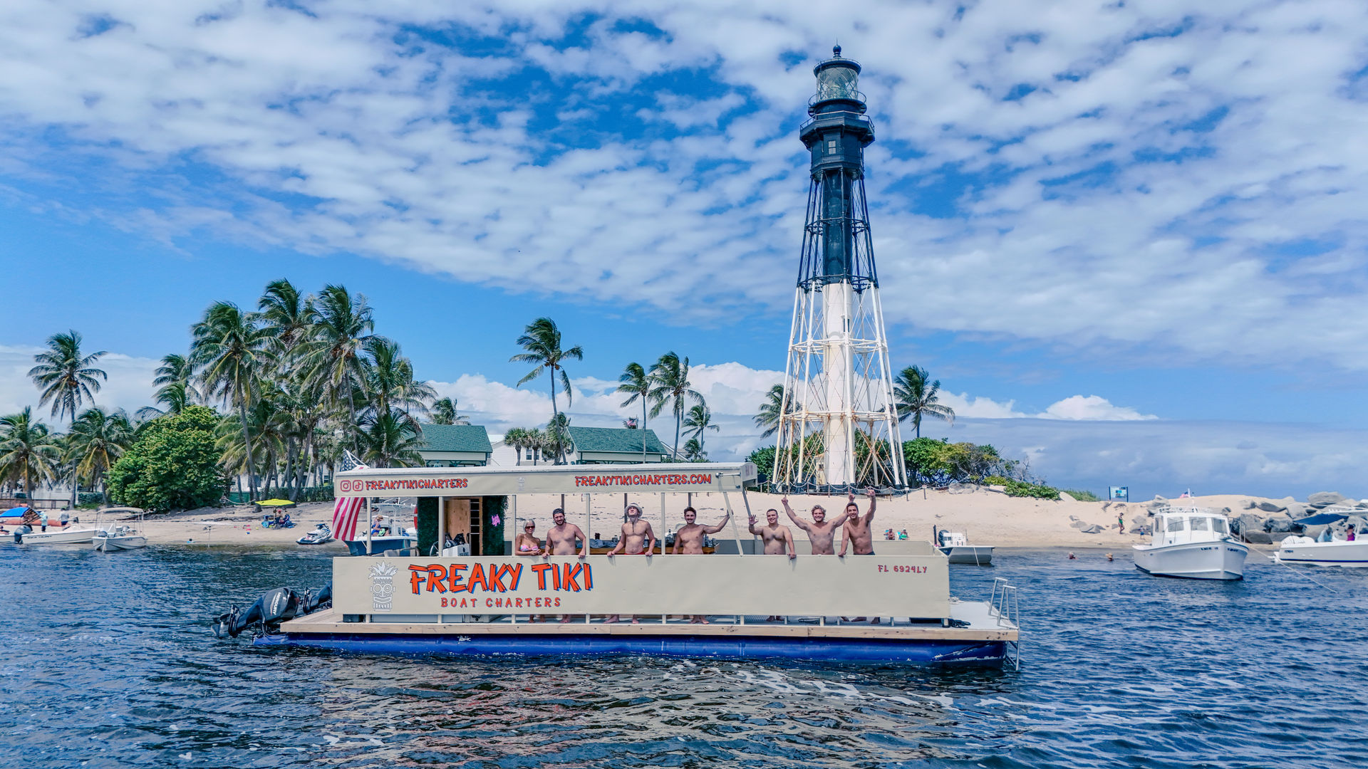 Party tiki boat with people waving near a tall black-and-white lighthouse off a palm-lined Florida beach under bright blue skies