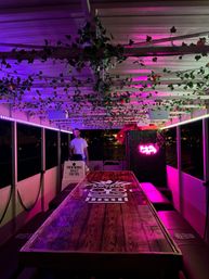 Nighttime party boat interior with long tiki‑style wooden table painted with a mask graphic, bench seating, pink and purple LED lighting, hanging faux vines on the ceiling, a neon sign, and a person standing near the helm with water visible outside.