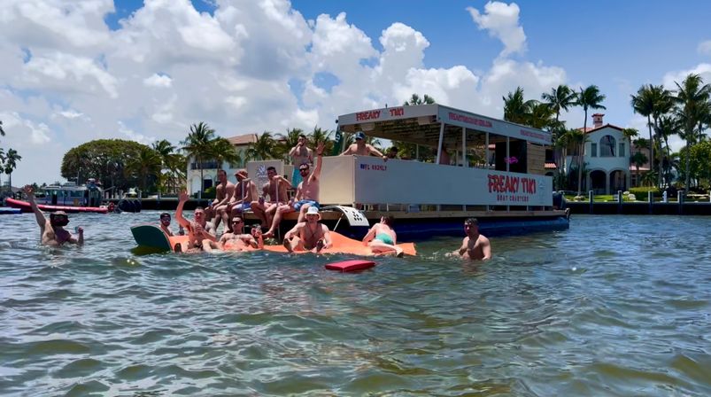 Group of people enjoying a sunny party boat and orange floating mats in a tropical waterfront inlet with palm trees and upscale homes in the background.