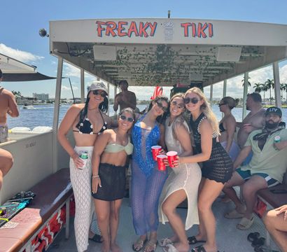 Smiling group of friends on a tiki-style pontoon boat at a sunny tropical waterfront with palm trees and red party cups