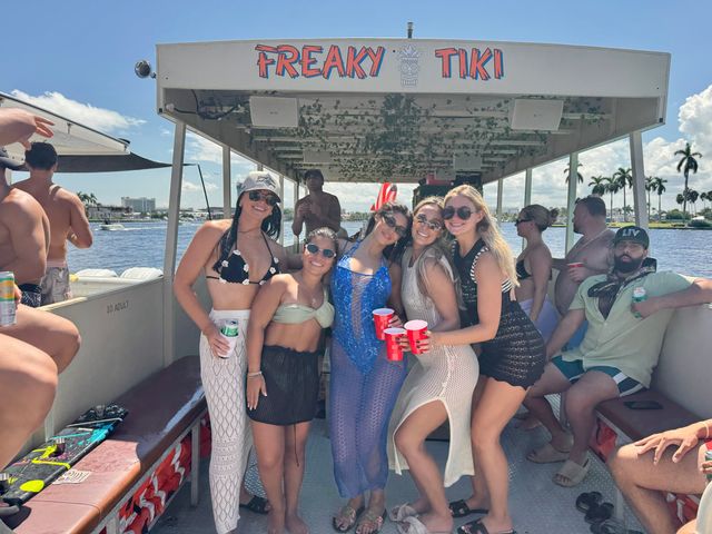 Smiling group of friends on a tiki-style pontoon boat at a sunny tropical waterfront with palm trees and red party cups
