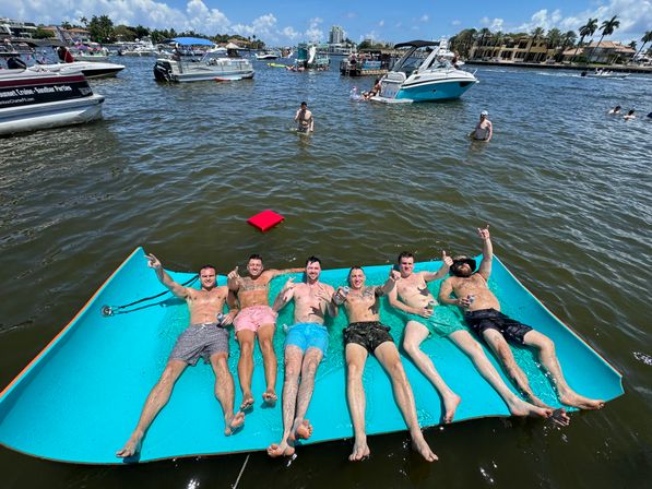 Six friends lounging on a turquoise floating mat in a sunny Florida inlet, surrounded by boats, yachts and a palm-lined waterfront