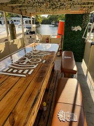 Sunlit waterfront canal boat interior with glossy wooden bar table featuring a tiki logo, brown leather bench seating, faux greenery wall and view of the canal