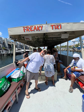 Sunny tiki-themed party boat at a marina, passengers socializing under a colorful canopy with docked yachts and waterfront buildings in the background.
