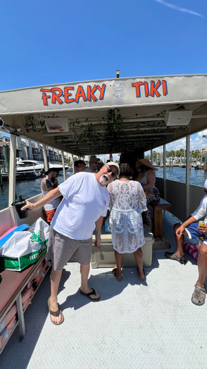 Sunny tiki-themed party boat at a marina, passengers socializing under a colorful canopy with docked yachts and waterfront buildings in the background.