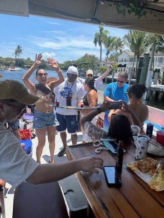 Sunny boat party on a canal — friends cheering on deck with palm trees and waterfront homes in the background.