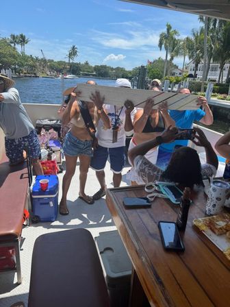 Boat party scene on a sunny tropical waterfront canal with palm trees and docks, a group of adults lining up to drink from a long wooden board while friends take photos