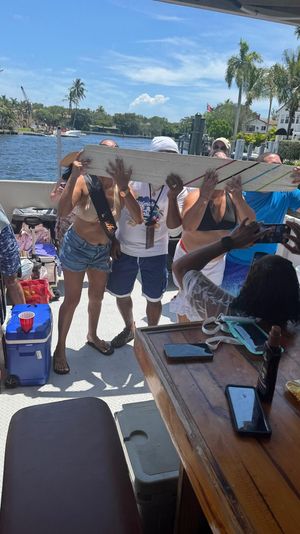 Boat party scene on a sunny tropical waterfront canal with palm trees and docks, a group of adults lining up to drink from a long wooden board while friends take photos
