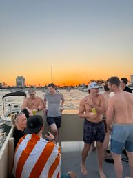 Group of people enjoying a party boat at golden sunset, palm-lined coastal marina with anchored yachts and distant buildings on the water