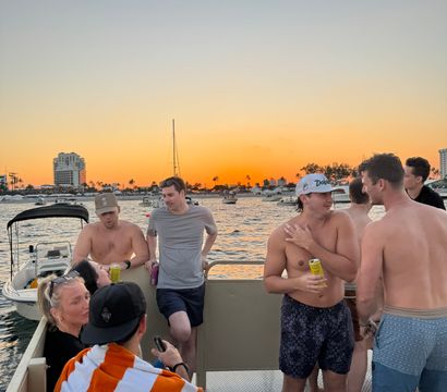 Group of people enjoying a party boat at golden sunset, palm-lined coastal marina with anchored yachts and distant buildings on the water