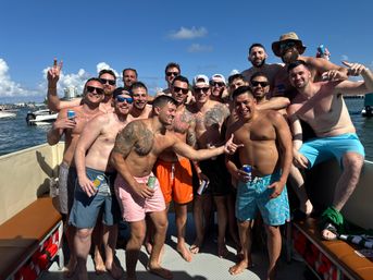 Group of shirtless men laughing and posing with drinks on a crowded party boat under a bright blue sky near a sunny coastal waterfront with boats in the background