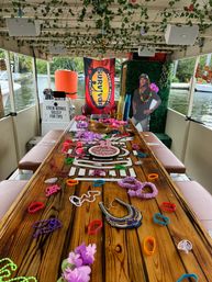 Party boat interior on a canal with a long wooden table covered in colorful leis, beads, wristbands and a '25' birthday cake, ivy-draped ceiling, a 'Survivor' banner and waterfront views.